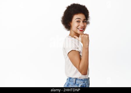 Portrait of smiling teen black woman, student pointing finger left, showing advertisement, standing in casual outfit over white background Stock Photo