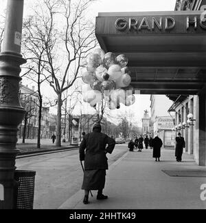 A woman selling balloons in Vienna. For hours the old lady of Vienna had tramped the cold wintry streets endeavouring to sell her wares flying from a string tied to her belt. At long last she made a sale, and a very contented customer walked away with his capture flying from a string attached to a button on his coast. The delight on his face and the delight on the old woman's face was worth the wait. Vienna, Austria. February 1954. Stock Photo