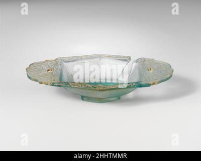 Glass dish ca. A.D. 100–150 Roman Translucent light blue green.Plain, rounded, horizontal rim; hexagonal body with straight sides tapering downward; integral hexagonal base, with ground flat edge; small flat bottom; two large integral handles, slightly upturned with rounded edge, each pierced with a central irregular star-shaped hole at base next to rim, and carved into a palmette shape with round and pointed projections. On upper surface incised decoration running around rim, on bottom in recessed hexagonal panel, and on handles: on rim, a continuous tendril scroll with circular flowers decor Stock Photo