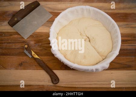 Freshly made sourdough bread in a proofing basket on a wood work surface with a lame and bench knife Stock Photo