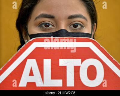 Young Mexican woman with beautiful brown eyes covers her face with disposable black face mask and red Mexican stop sign during corona pandemic. Stock Photo