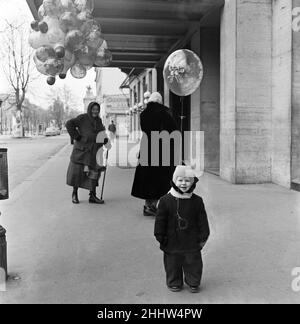 A woman selling balloons in Vienna. For hours the old lady of Vienna had tramped the cold wintry streets endeavouring to sell her wares flying from a string tied to her belt. At long last she made a sale, and a very contented customer walked away with his capture flying from a string attached to a button on his coast. The delight on his face and the delight on the old woman's face was worth the wait. Vienna, Austria. February 1954. Stock Photo