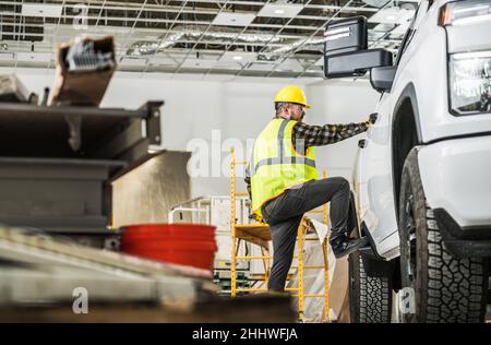 Caucasian Construction Worker in His 30s Wearing Hardhat Loading His Pickup Cargo Bed with Tools Inside Newly Built Warehouse Building. Stock Photo