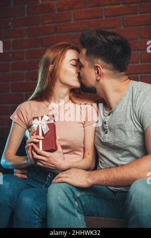 Woman kissing her boyfriend after receiving a birthday present while sitting on a couch posing over brick wall. Anniversary day. Stock Photo