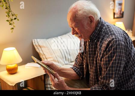 Senior man with grey hair and moustache looking at picture of his wife or children in wooden frame while sitting on bed Stock Photo