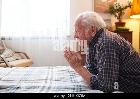 Old religious man with his hands put together in front of face leaning on bed while praying and praising Lord in the morning Stock Photo