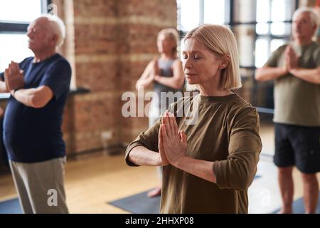Group of mature men and women keeping hands put together by chest while practicing yoga exercise in gym Stock Photo