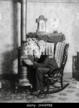 Elderly gentleman keeping warm close to stove. People still struggling to keep warm 100+ years later. Photograph from 1900. Stock Photo