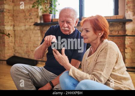 Elderly man pointing at smartphone screen during discussion with mature female at break after physical training in gym Stock Photo