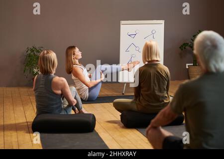 Young fitness instructor showing physical exercise to senior audience while sitting on mat with he legs bent in knees and raised Stock Photo