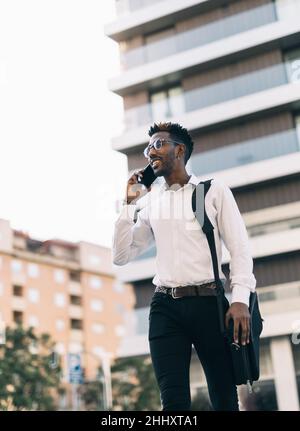 Happy and smiling young African American businessman walking across the street talking on his smart phone (vertical) Stock Photo