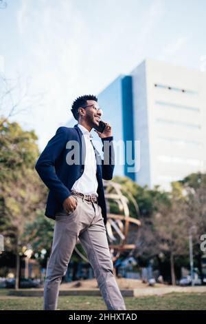 Smiling black man entrepreneur walking through a park while using his phone doing business Stock Photo