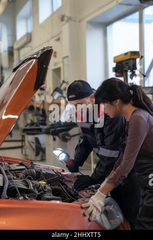 Two young intercultural mechanics in workwear bending over diesel engine of car while checking it before repairing Stock Photo