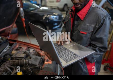 Part of repairman in workwear holding laptop while standing by car with open hood and consulting clients online in garage Stock Photo