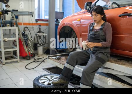 Young tired female worker of car maintenance service taking off gloves while sitting against car without wheel Stock Photo