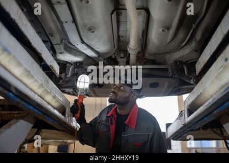 Serious young male technician of African ethnicity with lamp checking lower part of car while working in garage or workshop Stock Photo