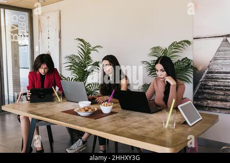 Trendy female colleagues using laptops and tablet while sitting at table and working on freelance project in modern cafeteria together Stock Photo