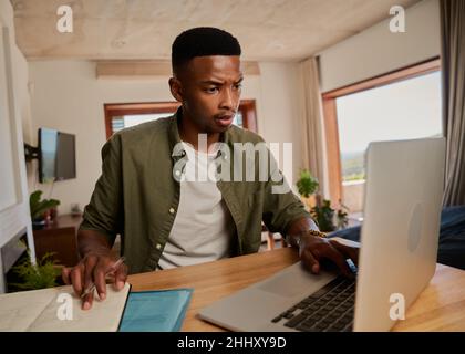 Young adult black male stressed, taking notes from laptop. Studying at home, sitting at modern styled kitchen counter. Stock Photo