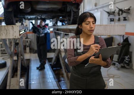 Young pretty female worker of car service making notes in document on clipboard against her male colleague repairing vehicle Stock Photo