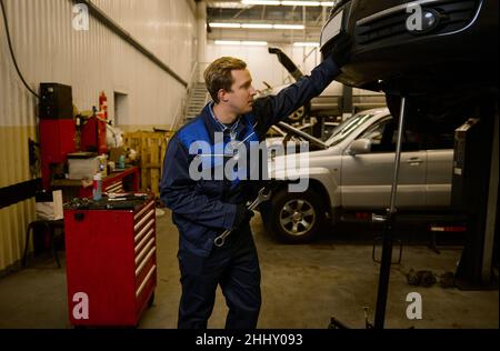 Handsome young mechanic in professional uniform, holding a wrench and working in auto service with lifted vehicle. Car repair and maintenance. Stock Photo