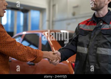 Handshake of young Hispanic female client and repairman in workwear standing against repaired car in garage or workshop Stock Photo
