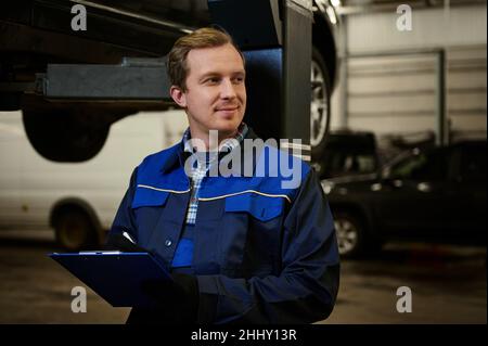 Portrait of auto mechanic, technician, car engineer in uniform making the checklist on clipboard for repairing the car lifted on a hoist in the repair Stock Photo