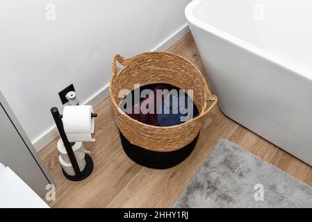 A wicker laundry basket with dirty clothes in the bathroom next to the bathtub. Stock Photo