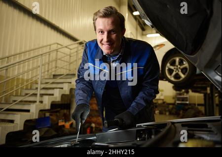 A smiling handsome Caucasian man, professional mechanic, auto technician repairing car engine at car service workshop. Stock Photo