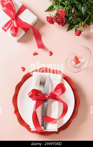 Valentines day table setting with gift, red roses, hearts on pink background. View from above. Valentine's invitation or greeting card. Vertical forma Stock Photo