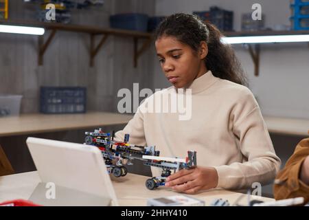 African girl sitting at the table in front of the tablet pc and making robot from constructor at science school Stock Photo