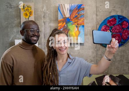 Multiethnic young couple making selfie portrait on mobile phone against the paintings in studio Stock Photo