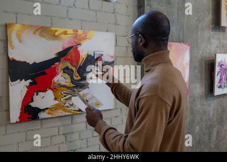 Rear view of African man using his mobile phone to make photo of artwork on the wall during exhibition Stock Photo