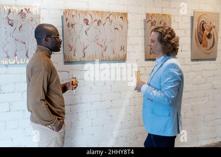 Two men standing near the wall with paintings drinking champagne and discussing modern art Stock Photo