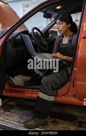 Young female technician with laptop sitting on front seat of car and answering online questions of clients Stock Photo