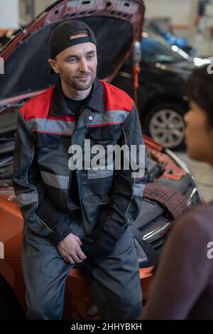 Young contemporary automechanic in workwear listening to female colleague while standing against open hood of car Stock Photo