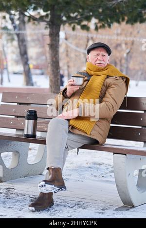 Elderly man in warm coat, scarf and cap having hot tea from thermos while sitting on bench in park on winter day Stock Photo