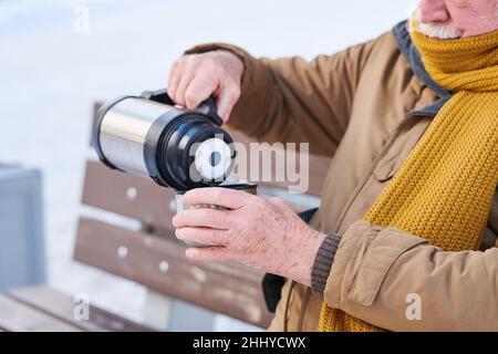 Contemporary aged man in warm coat and knitted scarf pouring hot tea from thermos into cup while sitting on bench on winter day Stock Photo