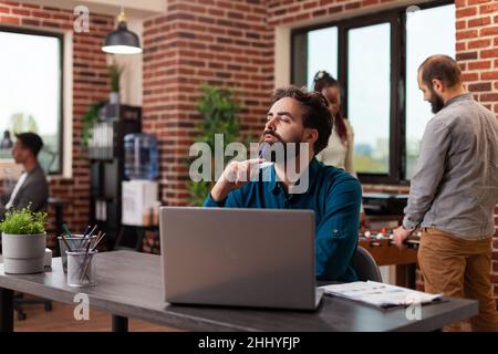 Pensive doubtful manager making serious expression while working at marketing project in startup office. Entrepreneur man thinking at company strategy. Diverse team planning business collaboration Stock Photo