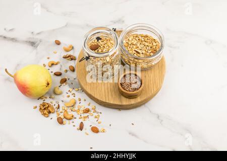 two different glass jars with walnut granola stand on a cutting round board. a marble background with scattered nuts on it and a ripe pear Stock Photo