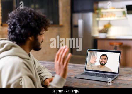Cheerful attractive middle eastern guy is using laptop for video communication, while sitting at the table in modern flat and waving into webcam. Talking online concept Stock Photo