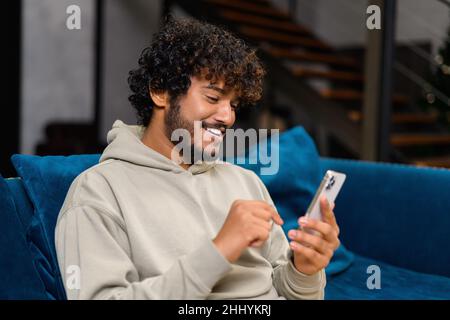 Focused bearded Indian man sitting on sofa at home and holding smartphone. Hispanic guy reading news online, spend leisure time in social networks. Dark-haired arab male texting and chatting online Stock Photo