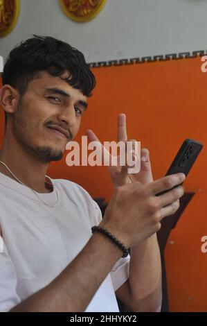 Closeup of a happy young guy looking at camera with showing peace sign with his hand while using mobile phone during sitting indoors Stock Photo