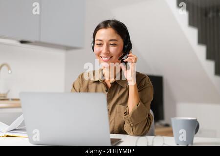 Young cheerful woman using headset for online talking sitting at the desk at home office. Pleasant female operator looking at the screen. Remote work concept Stock Photo