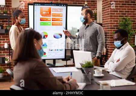 Entrepreneurs pointing at management presentation explaining financial strategy working in startup company office. Businessteam with medical face mask against covid19 brainstorming project ideas Stock Photo