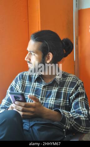 Selective focus of a good looking bearded man with hair bun, looking sideways during using his phone while sitting indoors Stock Photo