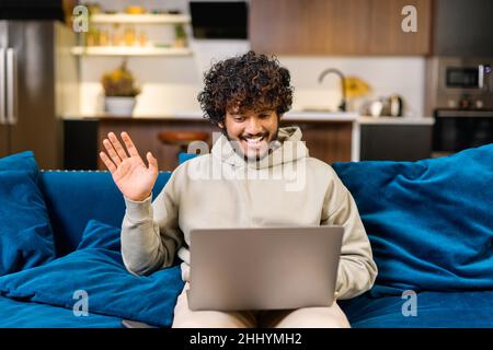 Video call. Cheerful attractive middle eastern guy is using laptop for video communication, while sitting at the sofa in modern flat and waving into webcam. Talking online concept Stock Photo