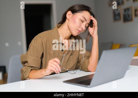 Exhausted multiracial entrepreneur businesswoman or student closed her eyes touching massaging her eyes, suffering from severe headache migraine. Tired young woman overwhelmed with computer work Stock Photo