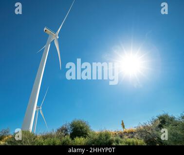 Huge wind turbine and tiny human silhouette on top of a hill against blue sky and shining sun, with lens flare Stock Photo