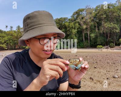 Happy asian male traveler caught and holds blue swimmer crab in hand on the beach with forest background. Wildlife, Adventure travel concept. Endau, M Stock Photo