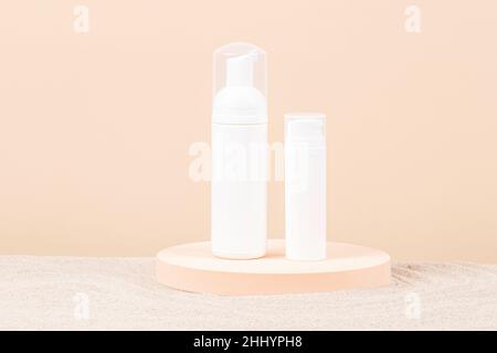 Two white blank plastic packaging cosmetic pump bottles for mousse foam cream and lotion stand on round podium, beach sand and beige background. Front Stock Photo